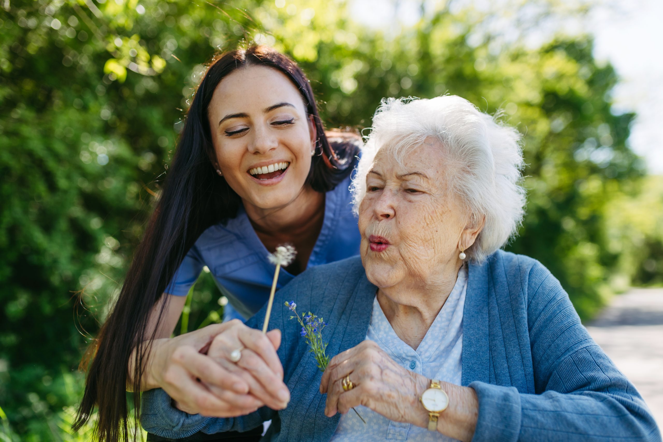 Female caregiver and senior woman enjoying outdoor time Caregiver and senior woman in wheelchair holding dandelion, picking wild flowers. Nurse and elderly woman enjoying a warm day outside their assisted living home.